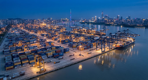 High-angle aerial photograph of deepwater shipping terminal in Guangzhou port loaded with blue and gold freight containers at blue hour with industrial gantry cranes reflecting in the calm ocean water